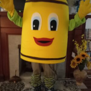 A person in a cheerful, yellow crayon mascot costume with big eyes and a smiling mouth poses indoors. The costume features brown stripes and colorful shoes.