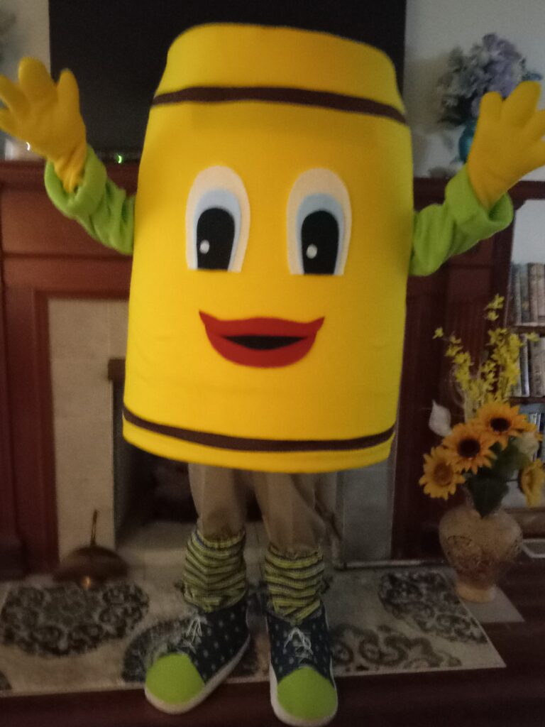 A person in a cheerful, yellow crayon mascot costume with big eyes and a smiling mouth poses indoors. The costume features brown stripes and colorful shoes.