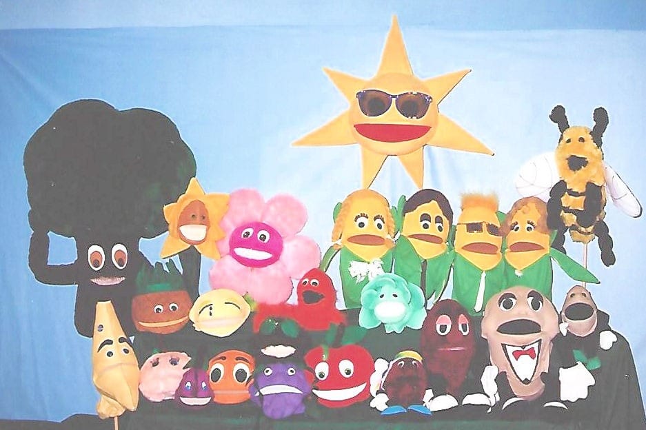 Colorful puppet characters, including a sun, tree, and various fruits and flowers, displayed against a light blue background, showcasing creativity and playfulness.