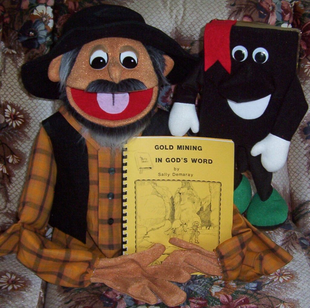 A cheerful puppet in a yellow plaid shirt and hat holds a book titled "Gold Mining in God's Word." A smiling book character stands beside it.