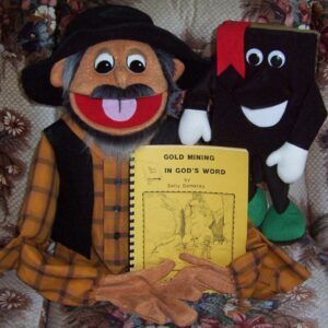 A cheerful puppet in a yellow plaid shirt and hat holds a book titled "Gold Mining in God's Word." A smiling book character stands beside it.